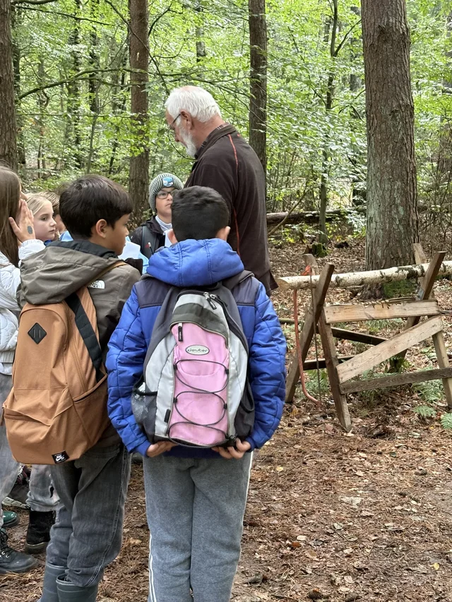Kinder mit Rucksäcken hören an einer Waldstation zu.